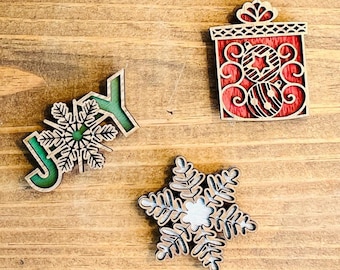 May include: Three wooden Christmas decorations: a red gift box with a white star, a green "JOY" with a snowflake, and a white snowflake.