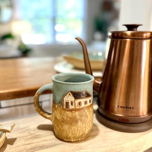 May include: A ceramic mug with a light blue upper section and a textured, speckled tan lower section. The mug features a small, hand-carved brown house design on the front. Beside it sits a metallic copper-colored electric kettle with a long spout.