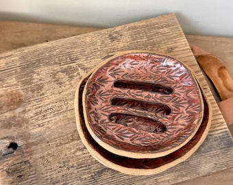 May include: A round, reddish-brown ceramic soap dish with a textured leaf pattern and three horizontal drainage slots, nested inside a matching plain ceramic base dish. The glaze has a glossy finish with darker accents in the recessed areas.