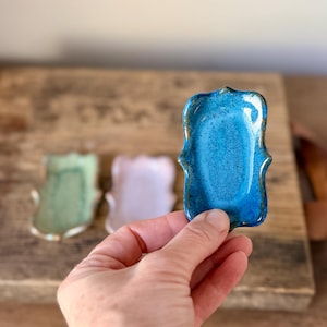 May include: A small, rectangular ceramic tray with wavy, scalloped edges and a glossy, vibrant blue glaze. The tray features a textured surface and dark, earthy brown trim along the outer rim, showcasing a handcrafted, organic appearance.