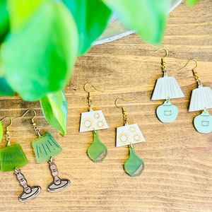 May include: Three pairs of green and white lampshade earrings. The lampshades are made of polymer clay and have a vintage style. The earrings are on a wooden surface.