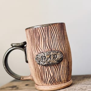 May include: A ceramic mug with a light brown, textured exterior resembling tree bark. An oval-shaped plaque is attached to the front, featuring the embossed text DAD. The mug has a dark, smooth handle and a matching dark rim, with a cylindrical body.