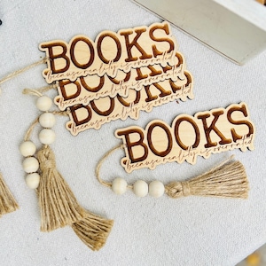 Puede incluir: Un conjunto de cuatro marcapáginas de madera con el texto "BOOKS because reality is overrated" grabado en cada uno. Los marcapáginas están unidos a borlas marrones y cuentas blancas.