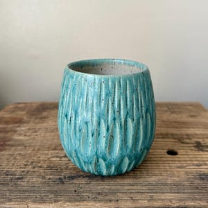 Puede incluir: Un vaso de cerámica turquesa hecho a mano con un diseño de ranuras verticales texturizadas. El pequeño recipiente redondeado tiene un interior moteado y se asienta sobre una superficie de madera. Ideal para bebidas.