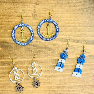 May include: A collection of nautical-themed earrings. Two pairs feature blue rope circles with gold anchors. Another pair showcases white lighthouse designs with wooden ship wheels. The final pair displays blue and white lighthouse silhouettes.