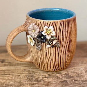May include: A ceramic mug with a textured tan exterior resembling tree bark, featuring a bright blue interior. The side is decorated with three-dimensional sculpted flowers in shades of white and grey, green leaves, and a small yellow and black bee.