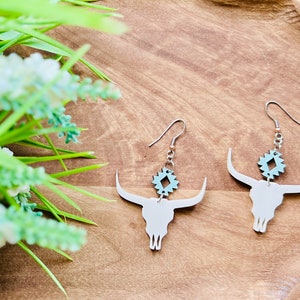 May include: A pair of silver dangle earrings featuring a white bull skull silhouette with a turquoise geometric charm hanging below.
