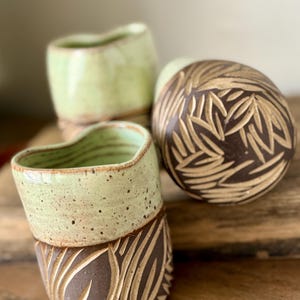 Puede incluir: Tres tazas de cerámica con un esmalte verde y un patrón de hojas marrones. Las tazas están hechas a mano y tienen un aspecto rústico.