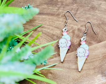 May include: A pair of dangle earrings featuring a cartoon ice cream cone with a smiling face and three scoops of ice cream in green, purple, and pink. The earrings are made of wood and have silver hooks.