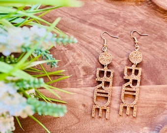 May include: Gold glitter 'BRIDE' earrings with a gold circle charm. The earrings are on a wooden surface.
