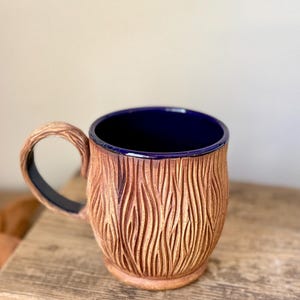 May include: A ceramic mug with a deep blue interior and a textured, light brown exterior featuring vertical, carved, wavy lines. The mug has a curved handle with a matching textured surface on the outside and a smooth, dark blue finish on the inside.