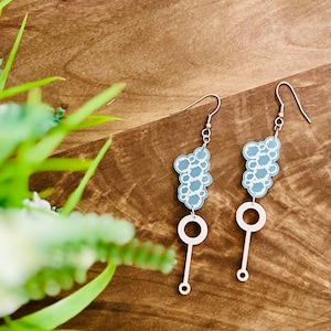 May include: A pair of silver dangle earrings with a blue bubble design and a silver circle charm. The earrings are on a wooden surface.