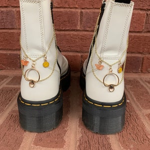 May include: Pair of white platform boots with black soles and yellow stitching. Gold chain accessories with mushroom, butterfly, and smiley face charms hang from the back of the boots. The boots have black pull tabs with yellow text.