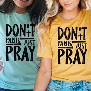 May include: Two women wearing t-shirts with the text "Don't Panic Just Pray" in black distressed lettering on a light blue and yellow background.