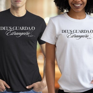 May include: Two t-shirts with the text "DEUS GUARDA O ESTRANGEIRO" in black lettering on a white background. The text is in a cursive font and includes the scripture reference "SL 146:9".