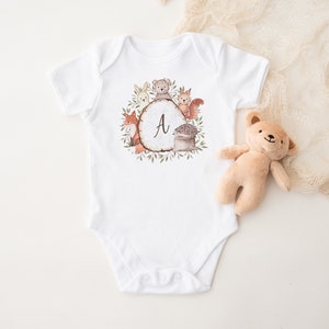 May include: White baby onesie with a woodland animal design. The design features a circle with the letter "A" in the center, surrounded by a fox, a bear, a rabbit, a squirrel, and a hedgehog. The animals are all in shades of brown, orange, and gray.