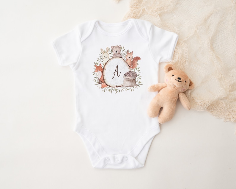 May include: White baby onesie with a woodland animal design. The design features a circle with the letter "A" in the center, surrounded by a fox, a bear, a rabbit, a squirrel, and a hedgehog. The animals are all in shades of brown, orange, and gray.