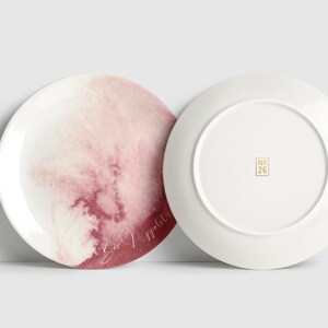 May include: Two white ceramic plates. One plate has a watercolour design in pink and red with the words "Bon Appetit" in cursive. The other plate is plain white with a gold "No. 26" stamp.