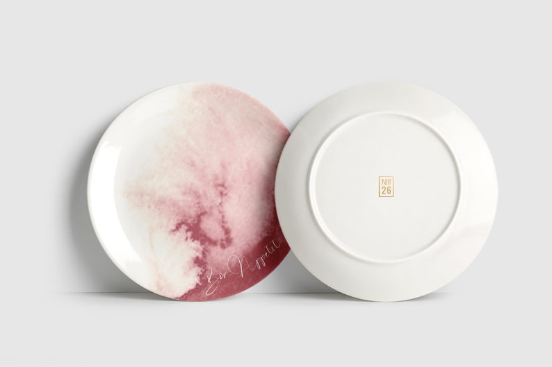 May include: Two white ceramic plates. One plate has a watercolour design in pink and red with the words "Bon Appetit" in cursive. The other plate is plain white with a gold "No. 26" stamp.