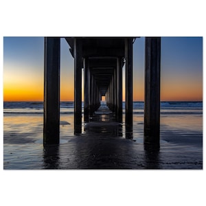 May include: A view through the pillars of a wooden pier, looking out to the sea at sunset. The water is calm and reflecting the orange and purple sky.