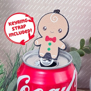 Peut inclure: Un porte-clés en forme d'homme en pain d'épices avec un nœud papillon rouge et des boutons verts, attaché à une canette rouge de Coca-Cola. Le porte-clés est livré avec une dragonne. Un bonhomme de neige est en arrière-plan.