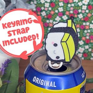 Pu&ograve; includere: Una lattina gialla di Twisted Tea, un t&egrave; freddo forte, con un portachiavi a forma di sushi bianco e nero in cima. Il portachiavi ha un viso da cartone animato carino ed &egrave; attaccato alla linguetta di apertura della lattina. La lattina &egrave; circondata da fiori rosa e viola.