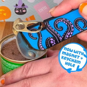 May include: A blue and black octopus-themed can opener is used to open a can of cat food. The can opener has a magnet and keychain hole. The background has Halloween-themed decorations.