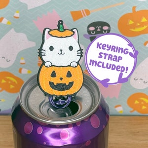 May include: A white cat with a pumpkin hat sits on top of a can of Bubly blackberry sparkling water. The cat is holding a small pumpkin with a jack-o-lantern face. The can is on a wooden surface. Keyring strap included.