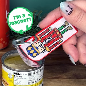 Puede incluir: Un imán rectangular con un diseño de soldado cascanueces, sostenido sobre una lata de comida abierta. El imán tiene el texto "I'm a magnet!". La etiqueta de la lata muestra información nutricional.