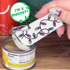 May include: A white magnet with a black outline of Christmas lights, with red, green, and yellow bulbs, is used to open a can of food. The text "I'm a magnet!" is in a green circle. A can of Bubly is in the background.