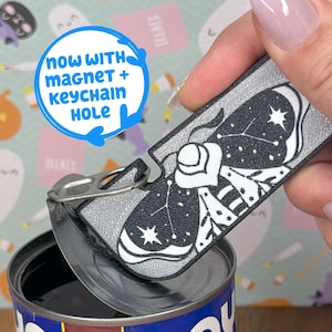 May include: Black and silver bottle opener with a constellation design, opening a can. The opener has a magnet and keychain hole. The text "NOW WITH MAGNET + KEYCHAIN HOLE" is in a blue circle.