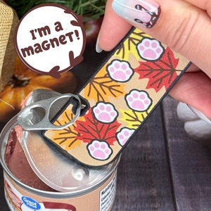 May include: A rectangular magnet with a fall-themed design featuring pink paw prints and colorful leaves. The magnet is attached to a can of potted meat. Text on the magnet says "I'm a magnet!"