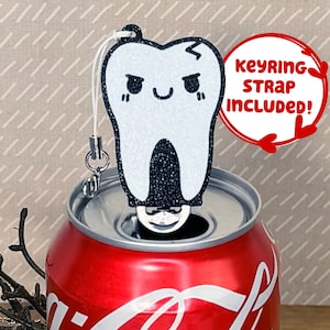 May include: A white glitter tooth-shaped can opener with a black glitter detail and a black angry face. The can opener is attached to a silver keyring with a white strap. The text "keyring strap included!" is written in red on a white circle.