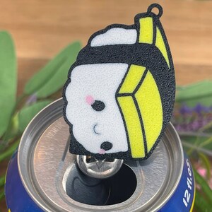 Pu&ograve; includere: Un ciondolo di sushi bianco e giallo con un involucro di alga nera. Il ciondolo &egrave; seduto sopra una lattina blu e gialla che dice "ORIGINAL" su di essa.