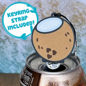 Op de afbeelding: Een bruine, kokosnootvormige flessenopener met een witte bovenkant en een zwarte sleutelkoord. De flessenopener ligt op een bruine frisdrankblikje met de tekst "No Caffeine" erop.