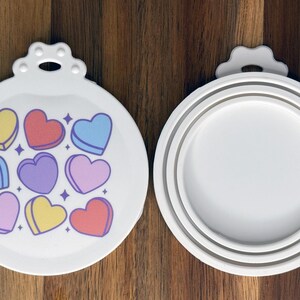 May include: Two white circular silicone lids with a tab at the top. The left lid features a pattern of nine colorful heart-shaped candies in yellow, red, blue, and purple with small star accents. The right lid shows the plain white underside.