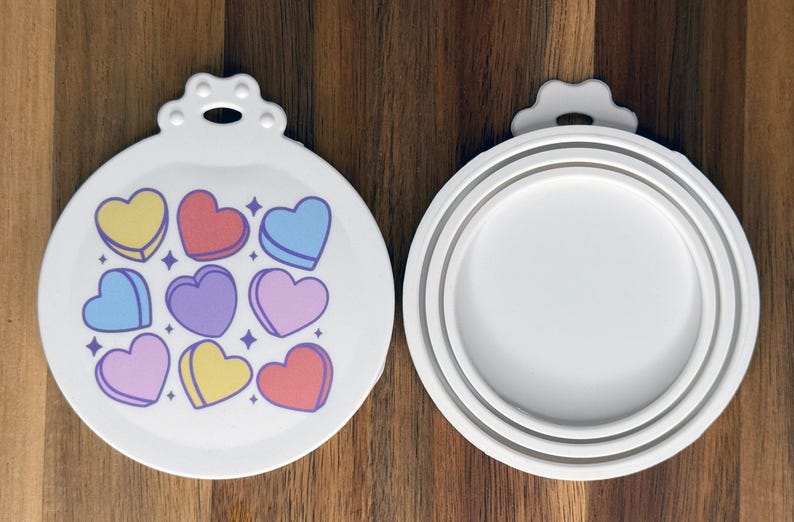 May include: Two white circular silicone lids with a tab at the top. The left lid features a pattern of nine colorful heart-shaped candies in yellow, red, blue, and purple with small star accents. The right lid shows the plain white underside.