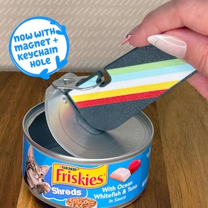May include: A cat food can with a blue and yellow label, opened to reveal the contents. A multi-colored, rectangular keychain with a magnet and keychain hole is being held above the can. Text on the can reads "Friskies Shreds".