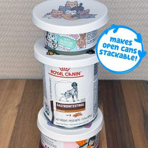 May include: A stack of three pet food cans with white plastic lids featuring a cartoon cat illustration. The middle can is Royal Canin Veterinary Gastrointestinal Low Fat dog food. A blue circular graphic with the text makes open cans stackable!
