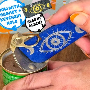 May include: A blue can opener with a silver eye design and crescent moons is opening a can of food. The text "NOW WITH MAGNET + KEYCHAIN HOLE" and "ALSO IN BLACK!" are in the image. A black version of the can opener is also shown.