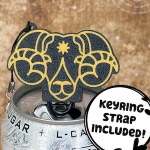 May include: A black and gold Aries zodiac sign keychain with a keyring strap. The keychain is attached to a soda can. The keychain has a textured surface and a decorative design.