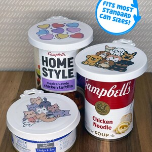 May include: Three white plastic reusable can lids with printed designs. One lid features colorful hearts, one shows a group of illustrated dogs, and one displays a pile of cartoon cats. A blue circular graphic states FITS MOST STANDARD CAN SIZES!