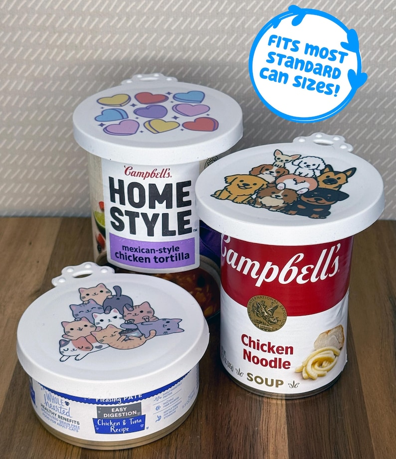 May include: Three white plastic reusable can lids with printed designs. One lid features colorful hearts, one shows a group of illustrated dogs, and one displays a pile of cartoon cats. A blue circular graphic states FITS MOST STANDARD CAN SIZES!