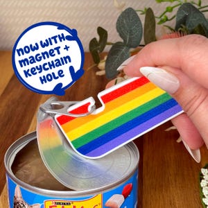 May include: A rainbow-colored can opener with a magnet and keychain hole is shown opening a can of cat food. The can opener features a rainbow design with red, orange, yellow, green, blue, and purple stripes. The text "NOW WITH MAGNET + KEYCHAIN HOLE" is in a blue circle.
