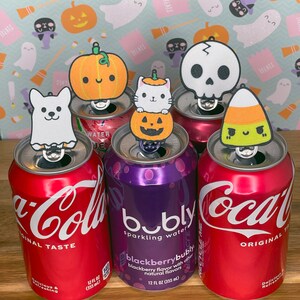 May include: Six Halloween-themed bottle toppers, including a ghost, pumpkin, skull, candy corn, and two pumpkins, are placed on top of various soda cans, including Coca-Cola, Bubly, and a generic brand.