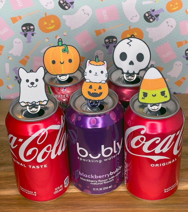 May include: Six Halloween-themed bottle toppers, including a ghost, pumpkin, skull, candy corn, and two pumpkins, are placed on top of various soda cans, including Coca-Cola, Bubly, and a generic brand.