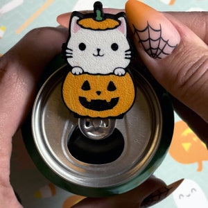 May include: A white cat wearing a pumpkin hat sits inside a carved pumpkin. The pumpkin has a black jack-o'-lantern face. The pumpkin is attached to a green can with a silver pull tab.