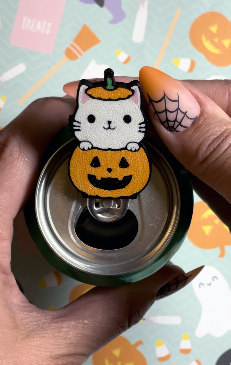 May include: A white cat wearing a pumpkin hat sits inside a carved pumpkin. The pumpkin has a black jack-o'-lantern face. The pumpkin is attached to a green can with a silver pull tab.