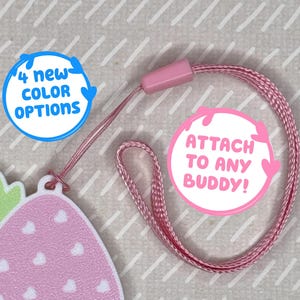 Puede incluir: Un charm en forma de fresa rosa y blanca con un tallo verde y corazones blancos. El charm está unido a un cordón rosa con un clip de plástico rosa. El cordón tiene un círculo rosa con el texto "ATTACH TO ANY BUDDY!" y un círculo azul con el texto "4 NEW COLOR OPTIONS".