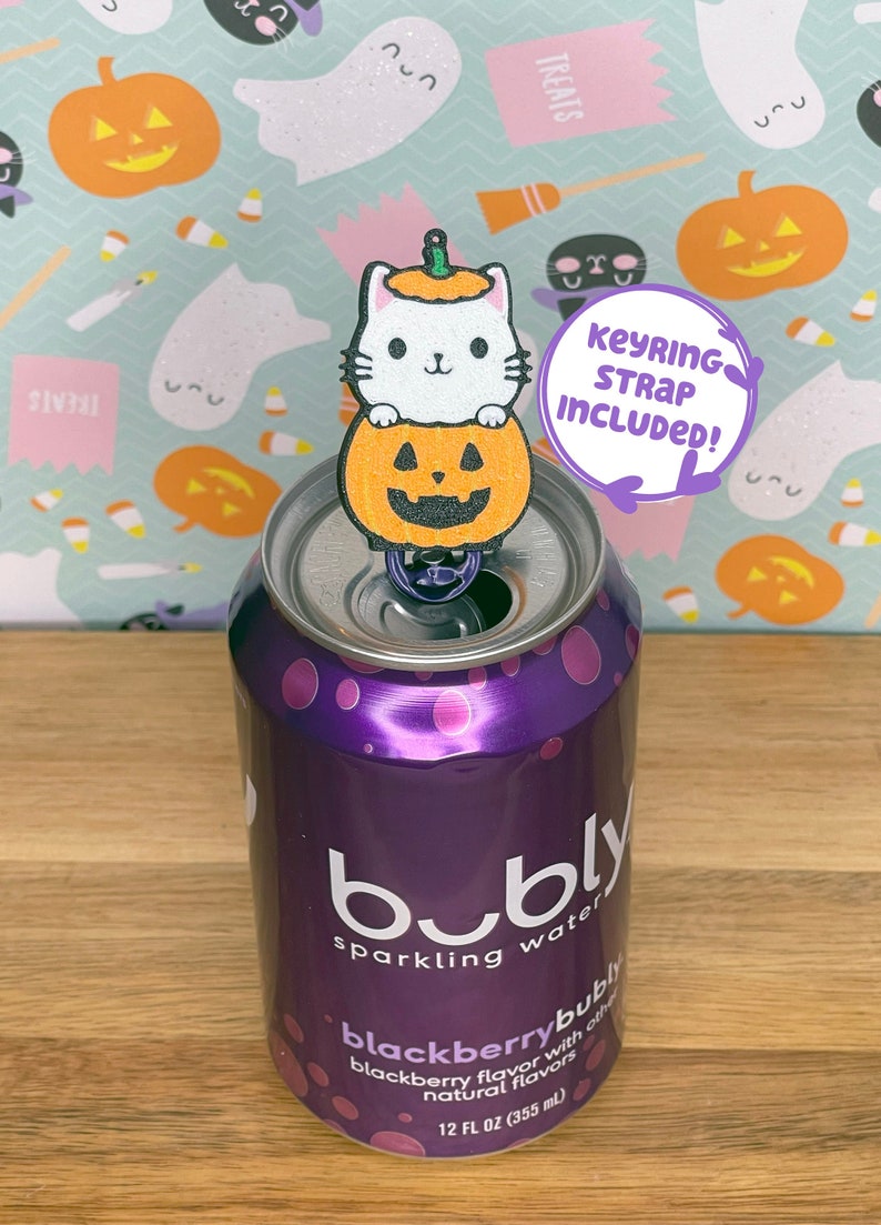 May include: A white cat with a pumpkin hat sits on top of a can of Bubly blackberry sparkling water. The cat is holding a small pumpkin with a jack-o-lantern face. The can is on a wooden surface. Keyring strap included.